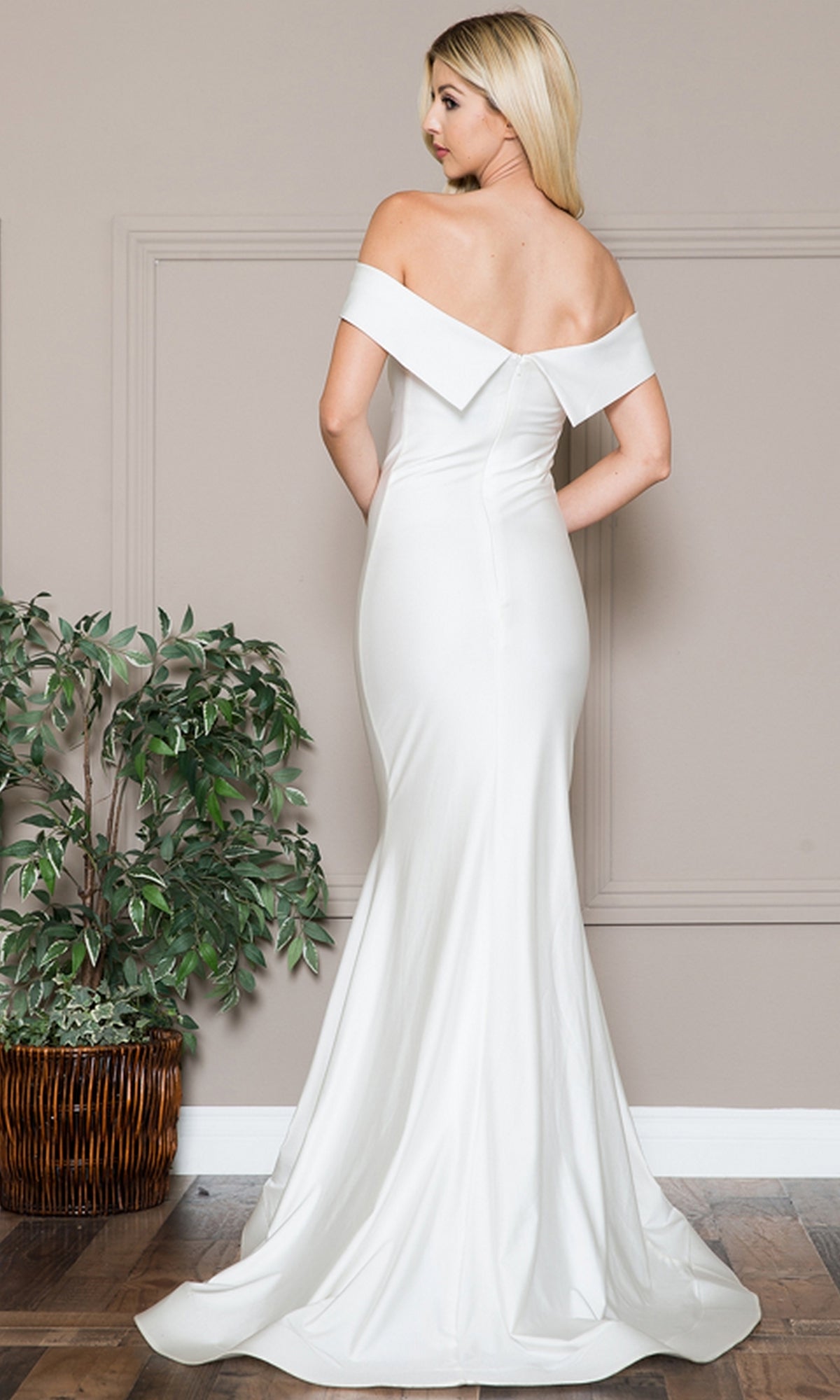 A woman stands indoors beside a potted plant, wearing the Sleek Simple Off-the-Shoulder Long Prom Dress 373, a floor-length white gown with a fitted bodice and flared hem, facing away from the camera.